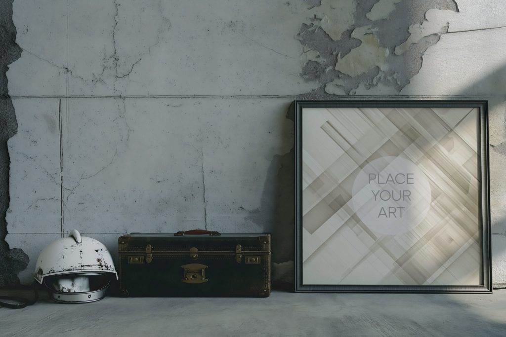 Large square wall art mockup leaning on a raw cement floor against a distressed concrete wall, styled with a vintage white motorcycle helmet and an antique dark leather trunk under harsh industrial shadows.