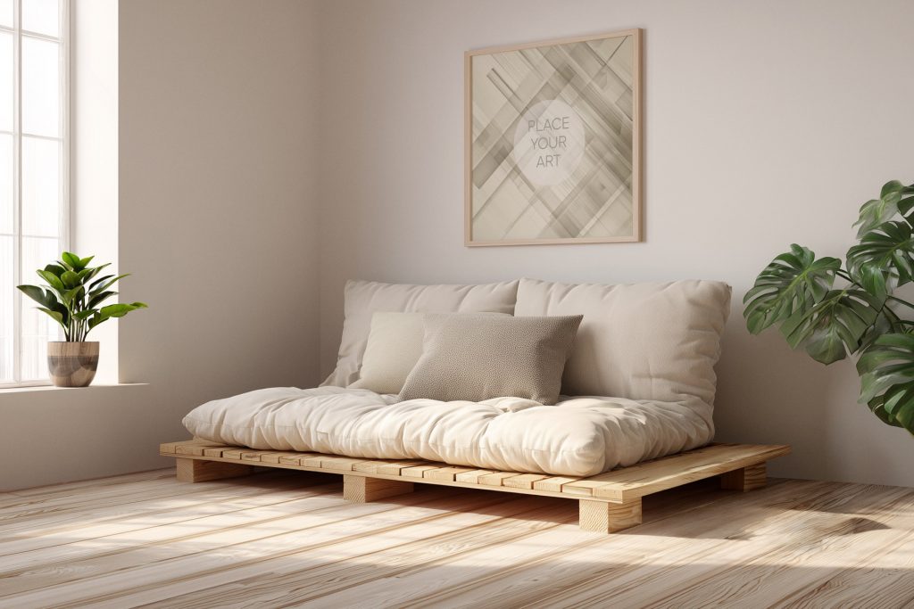 Square wooden frame mockup hanging above a low beige futon sofa on a wooden pallet frame with a large window and green plants.