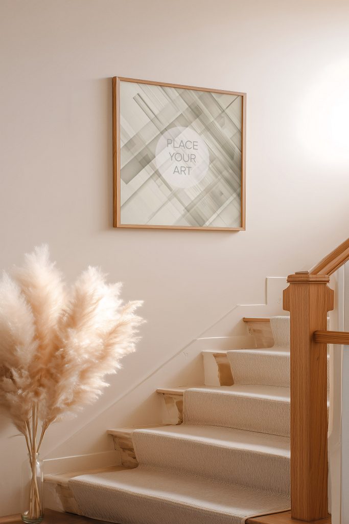 A square art frame mockup hanging on a neutral wall next to a staircase, styled with a glass vase of fluffy pampas grass in a sunlit interior.